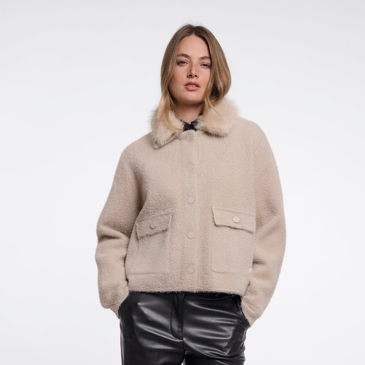 A model stands against a light gray studio background wearing a beige cropped shearling-style jacket with a large collar and flap pockets, paired with shiny black wide-leg pants and black shoes.
