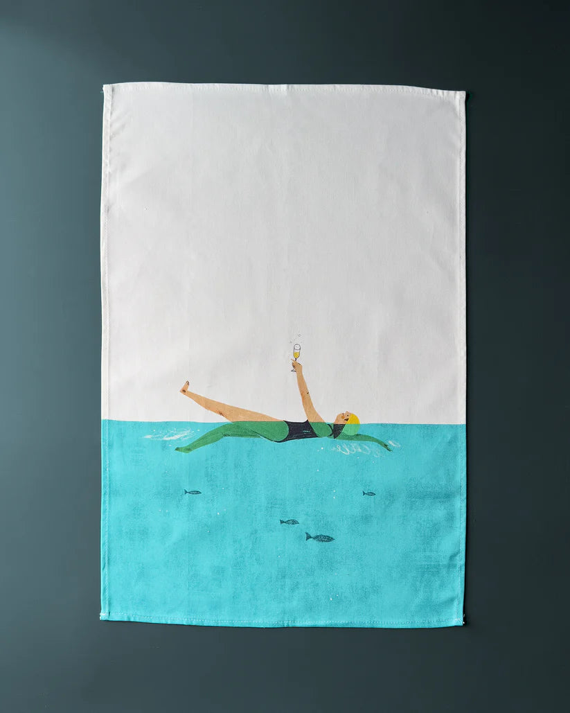 Tea towel featuring illustration of woman in teal swimsuit floating in turquoise water, holding champagne glass; small fish