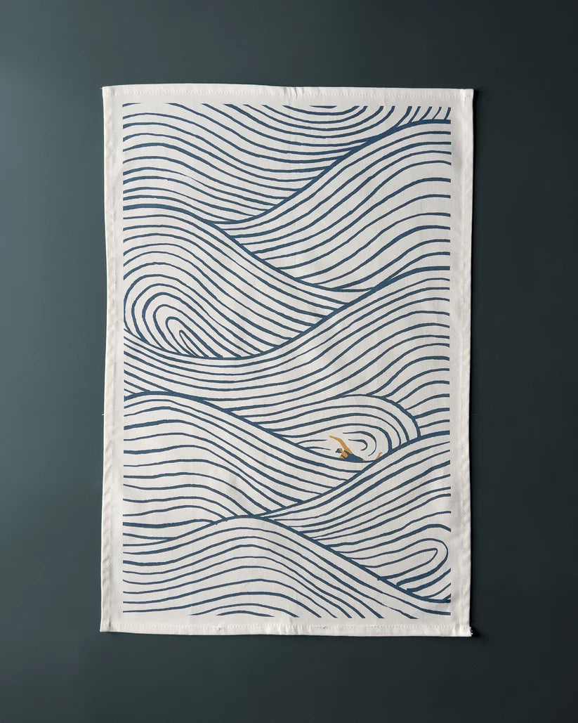 Tea towel with a stylized blue wave pattern on an off-white background.  A small gold detail is visible within the waves.