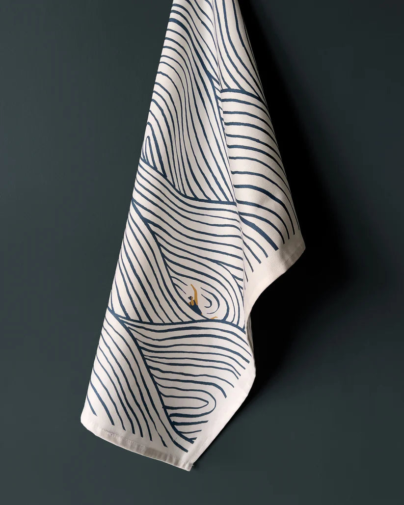 Cream-colored tea towel with a navy blue wave pattern and a small illustration of a swimmer.