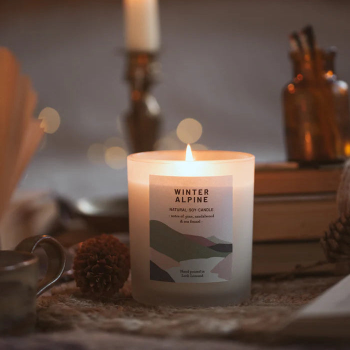 A lit frosted glass Winter Alpine soy candle with a minimalist label depicting abstract landscape.