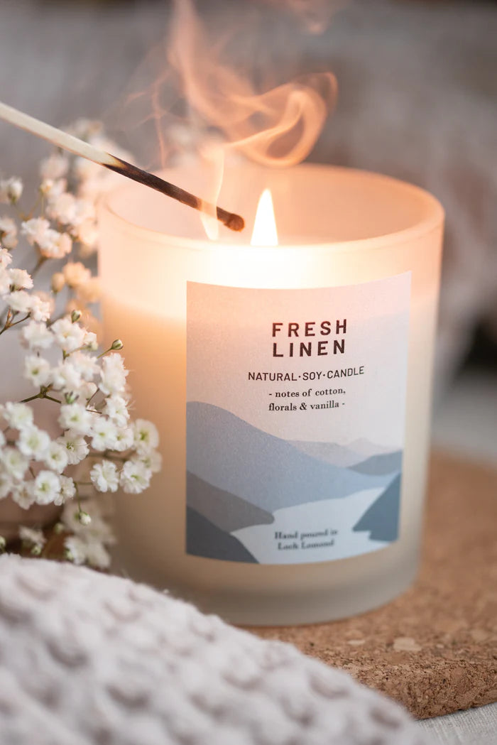 Lit natural soy candle in frosted glass jar.  Label reads "Fresh Linen," with scent notes of cotton, florals, and vanilla.