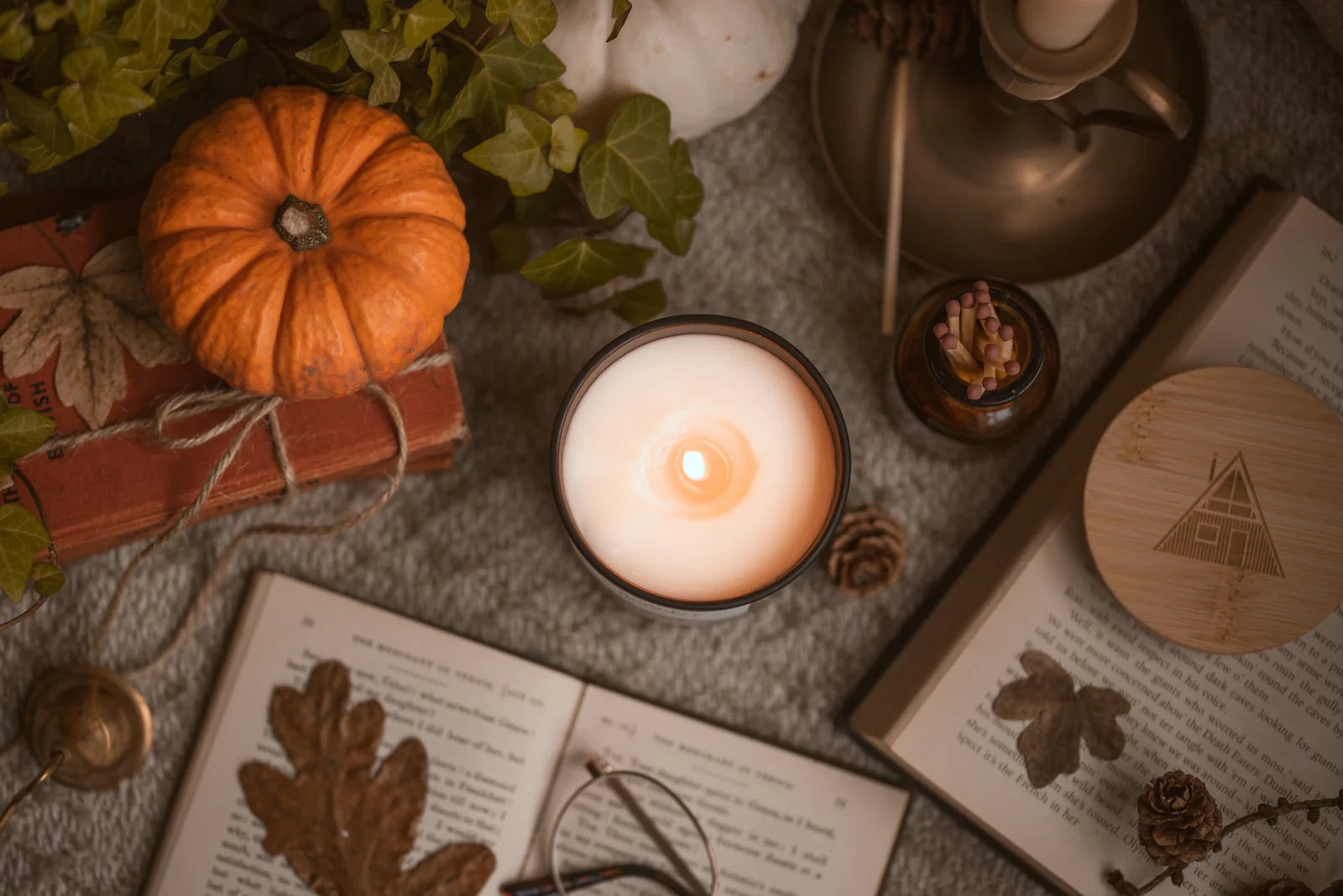 A lit candle in a dark-colored jar sits atop a textured surface, surrounded by autumnal items including a small pumpkin,