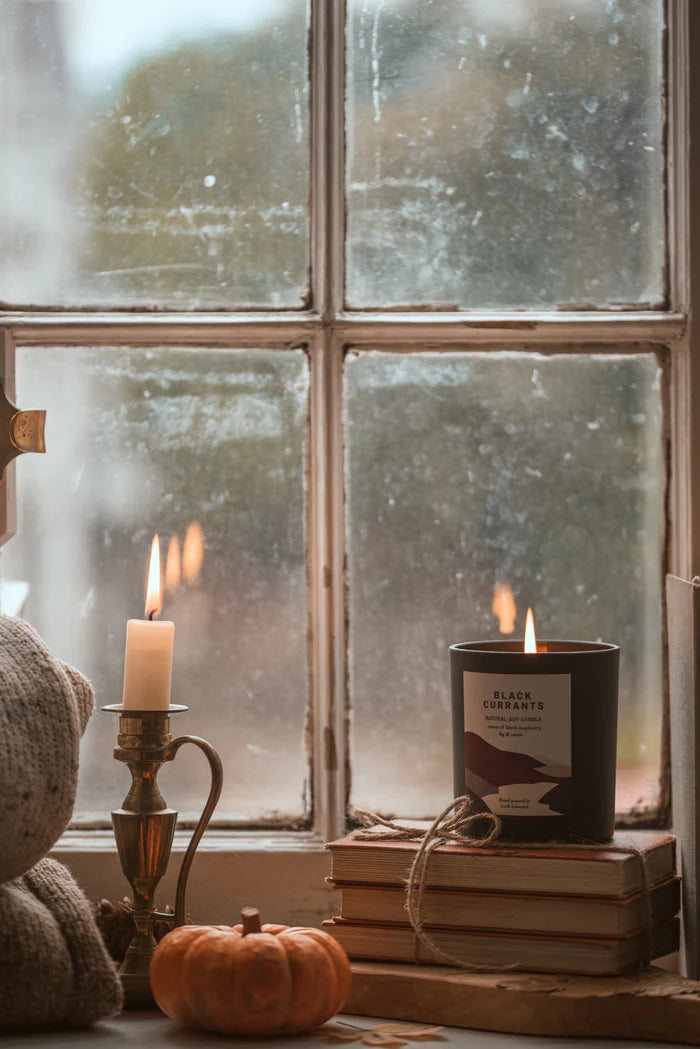 A lit Black Currants soy candle sits atop a stack of books near a lit candle in a brass candlestick holder.