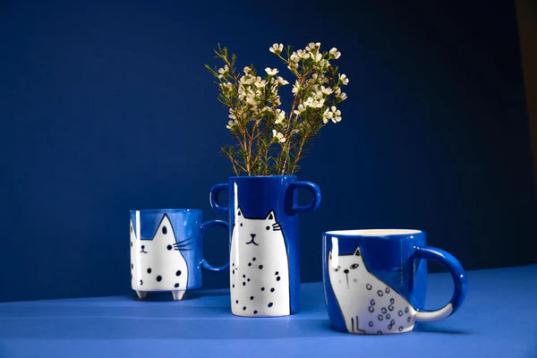 Blue ceramic mugs and vase with white cat designs.  The vase holds white flowers.