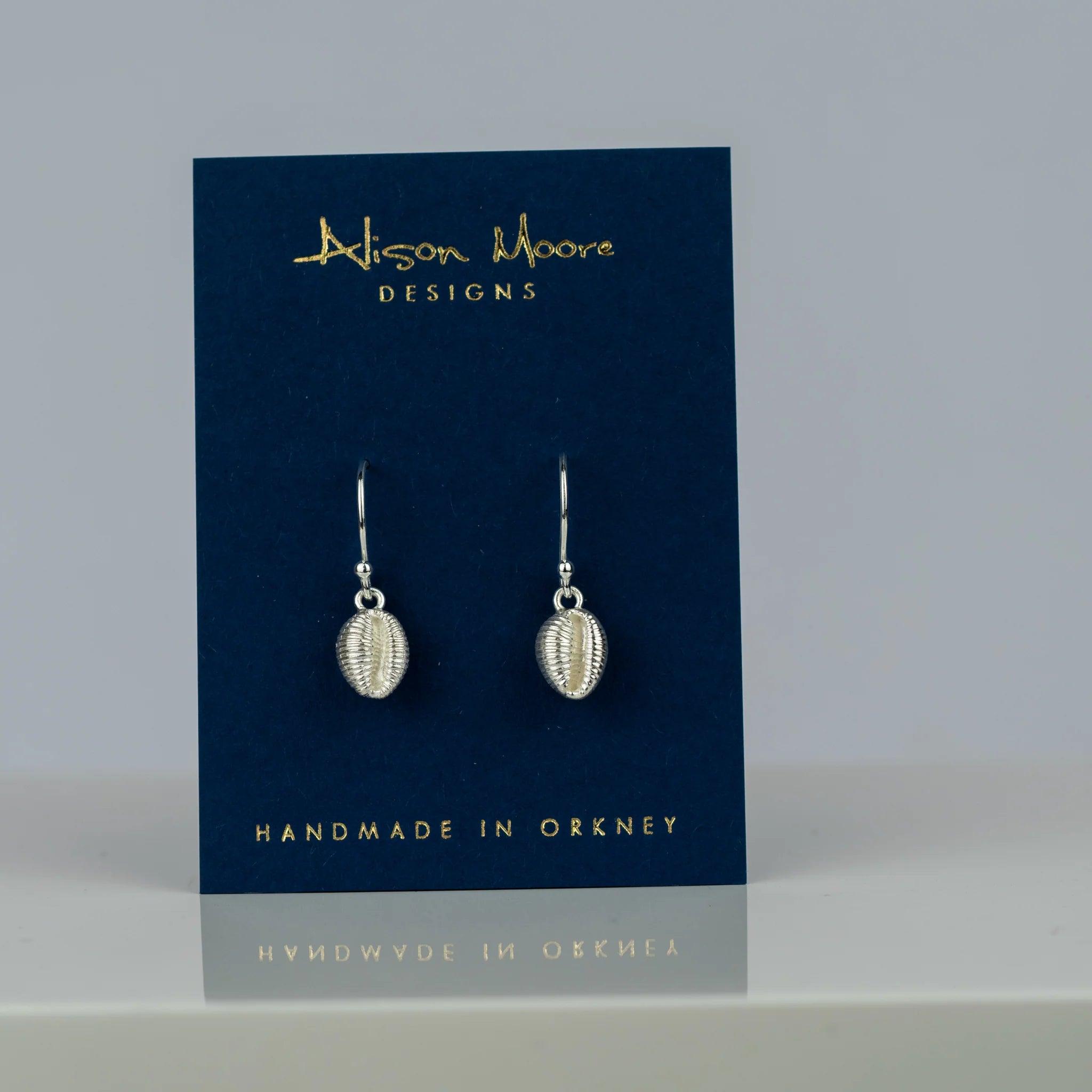 Pair of silver shell-shaped drop earrings on a navy blue display card.  Handmade in Orkney.