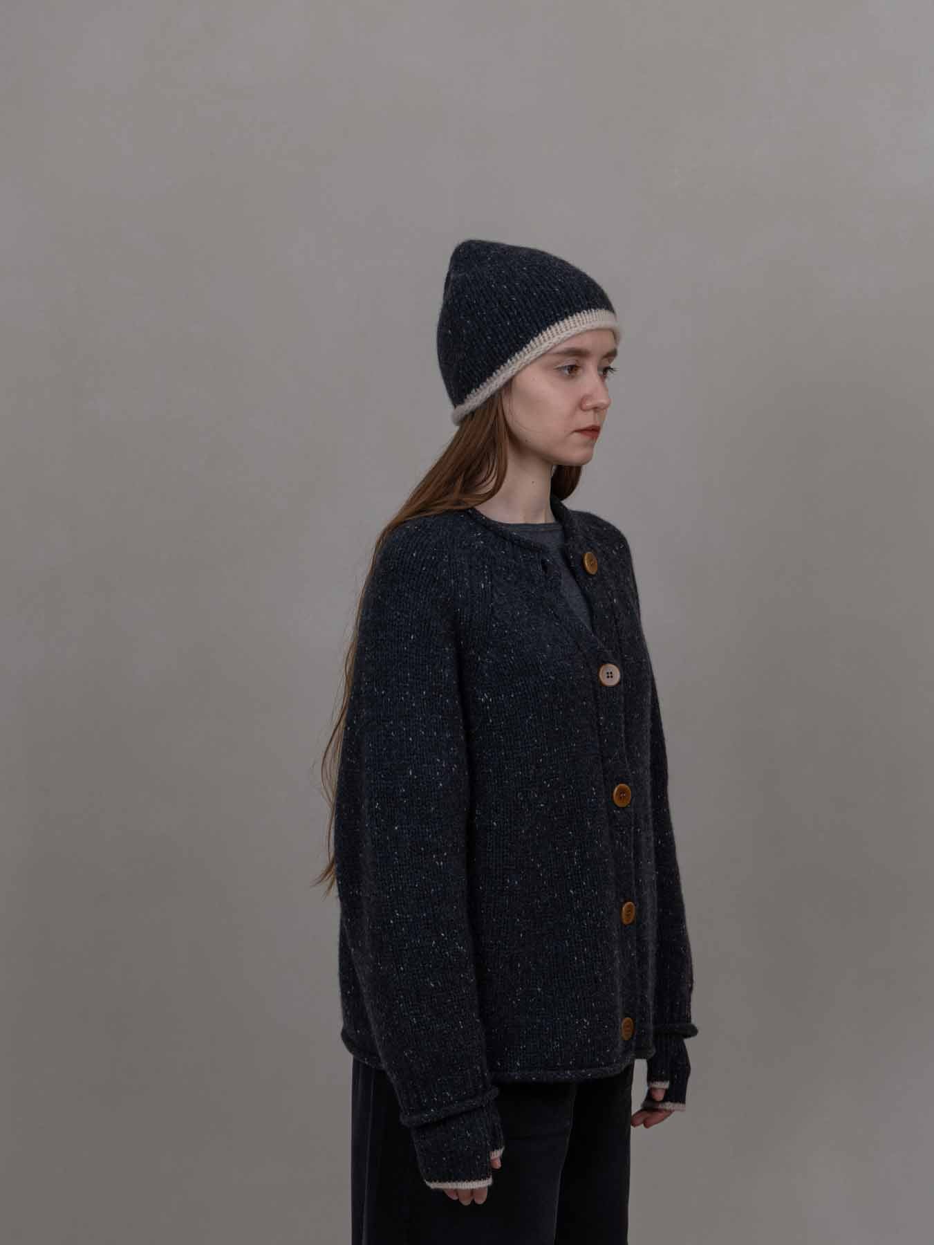 A woman wearing a dark navy textured knit cardigan with brown buttons and a matching dark knit beanie with a light beige trim, standing against a plain gray studio background.