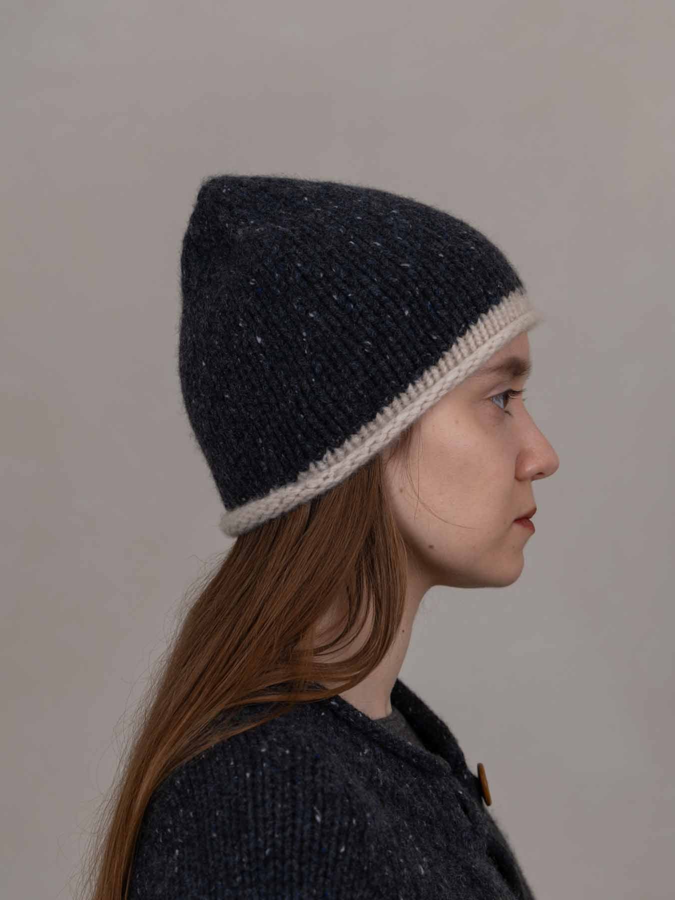 A side-profile portrait of a woman wearing a dark navy knit beanie with a contrasting cream-colored brim, set against a plain, warm gray background.