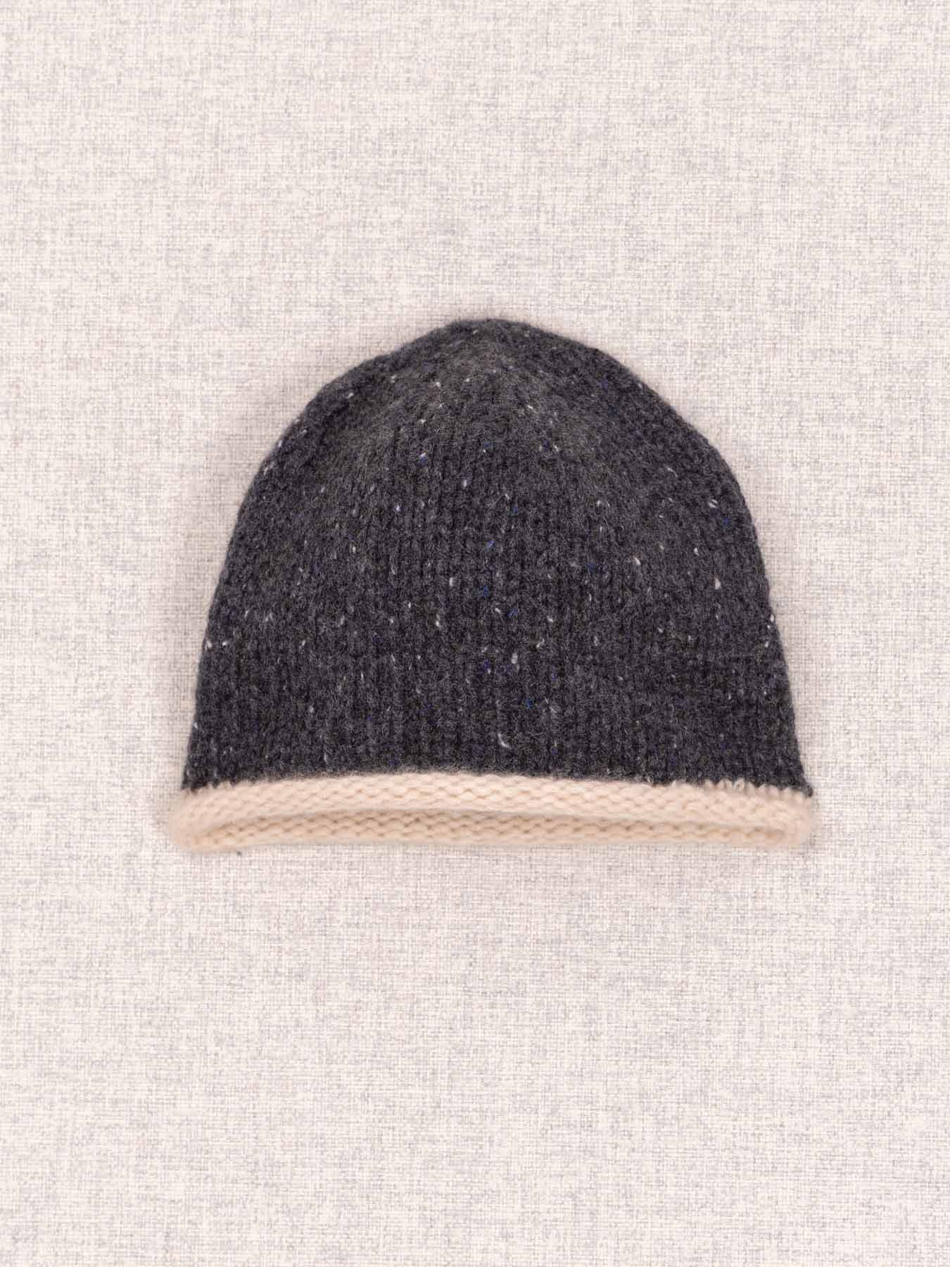 A dark charcoal gray knit beanie hat with a contrasting light beige ribbed cuff, displayed flat against a textured light beige fabric background.