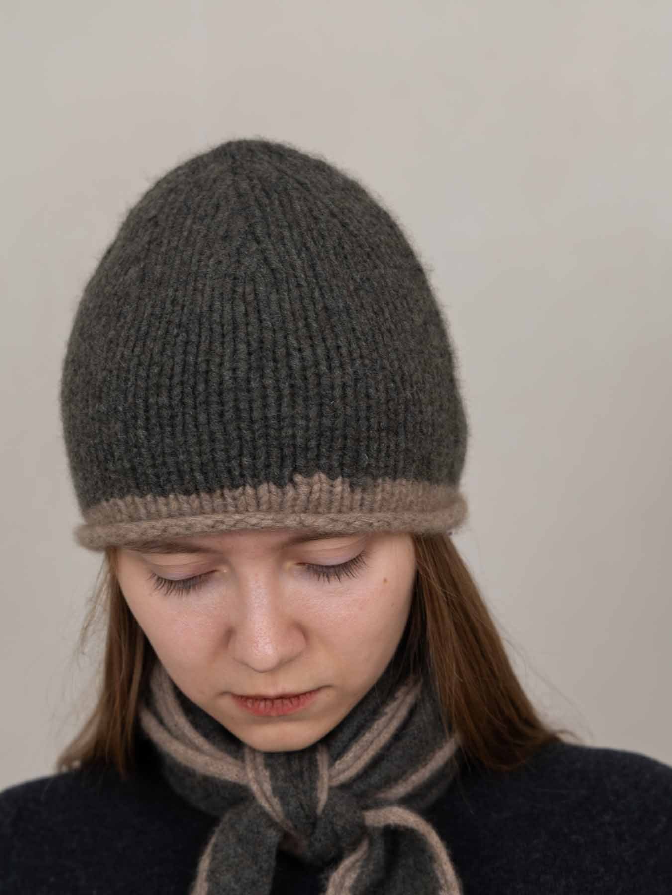A woman wearing a dark gray ribbed knit beanie with a contrasting light brown brim and a matching gray scarf, looking down against a plain light gray background.