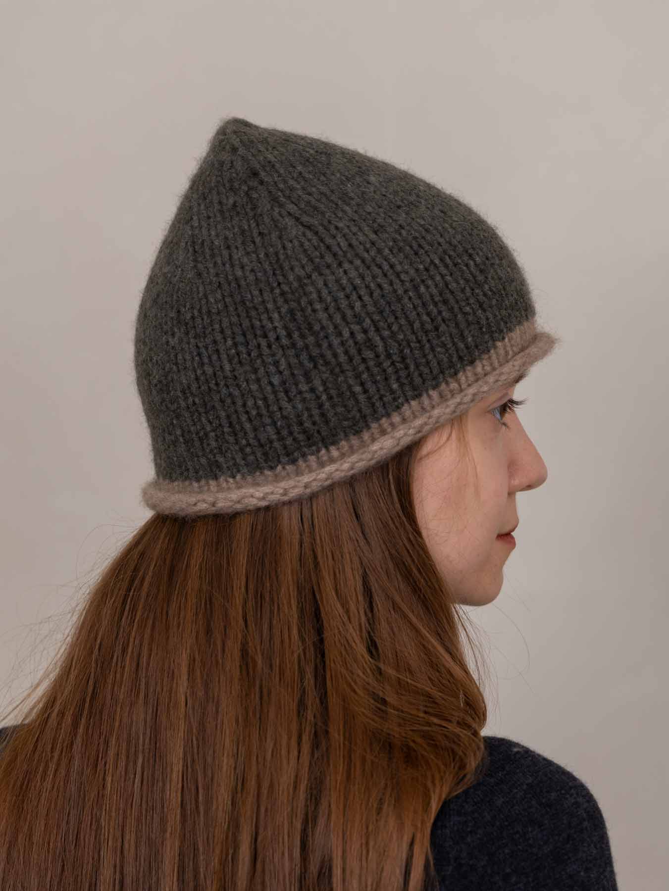 A woman in profile wearing a dark gray knit beanie with a contrasting light brown brim, set against a plain light gray background.