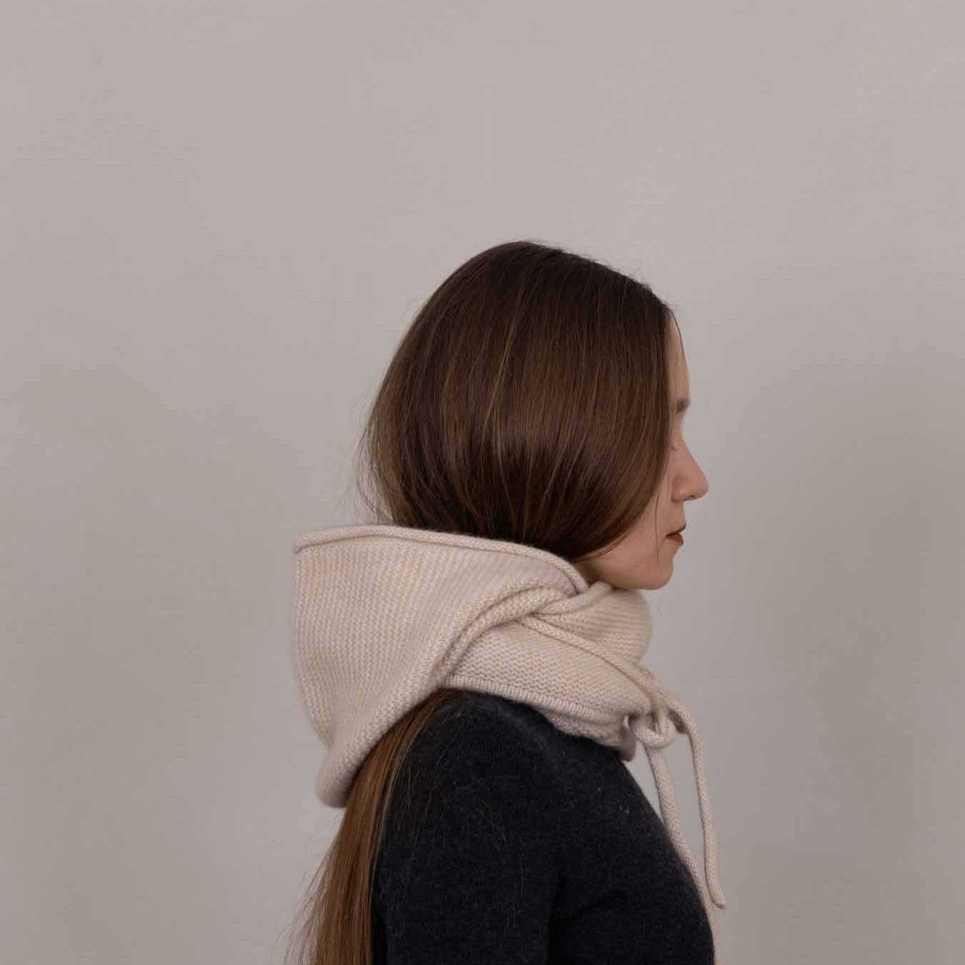A woman in profile wearing a cream-colored ribbed knit hooded scarf over a dark navy sweater, set against a plain light gray background.