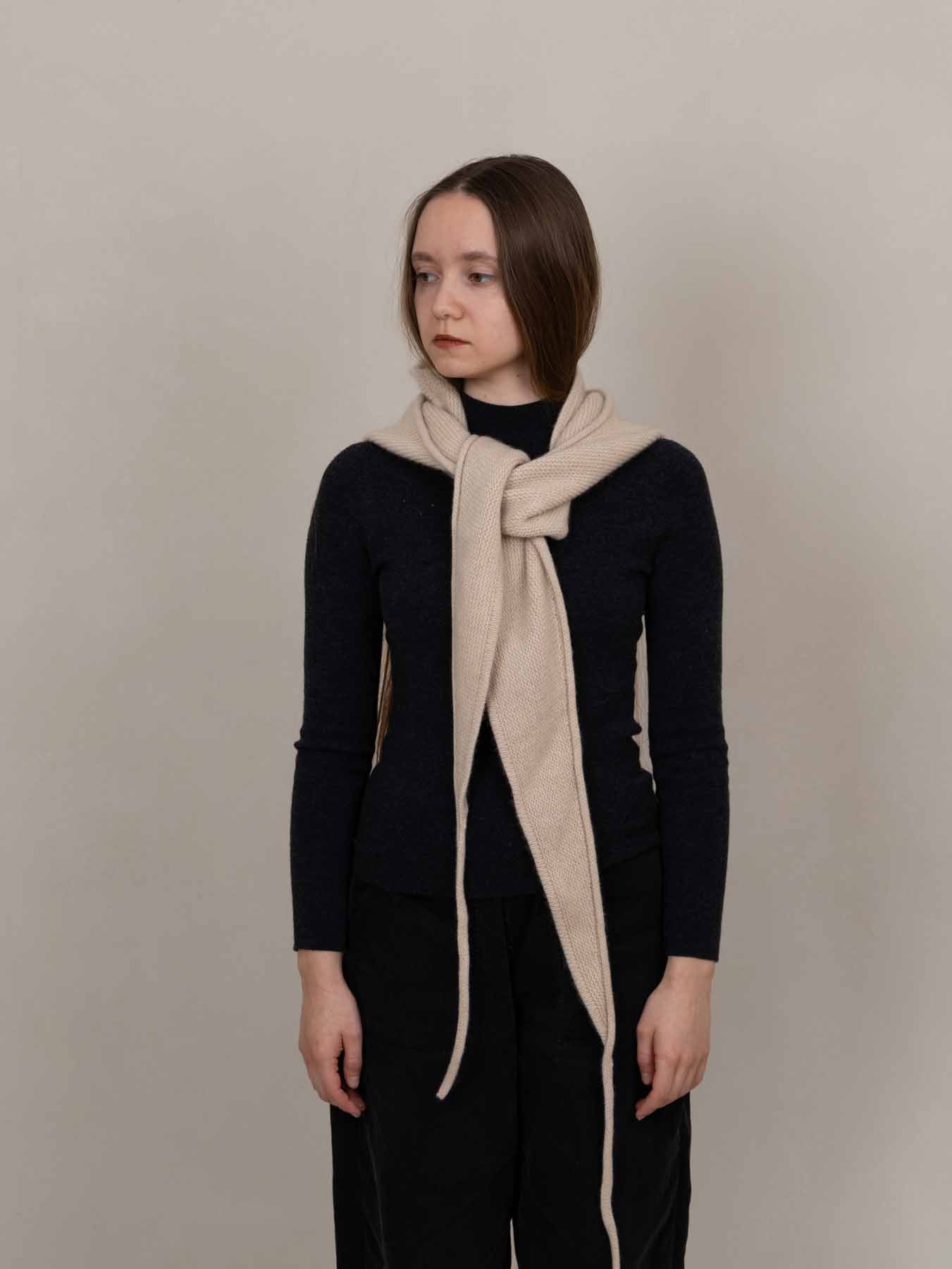 A woman wearing a dark navy long-sleeve top and black pants, styled with a thick cream-colored knit scarf wrapped around her neck, standing against a plain beige studio background.