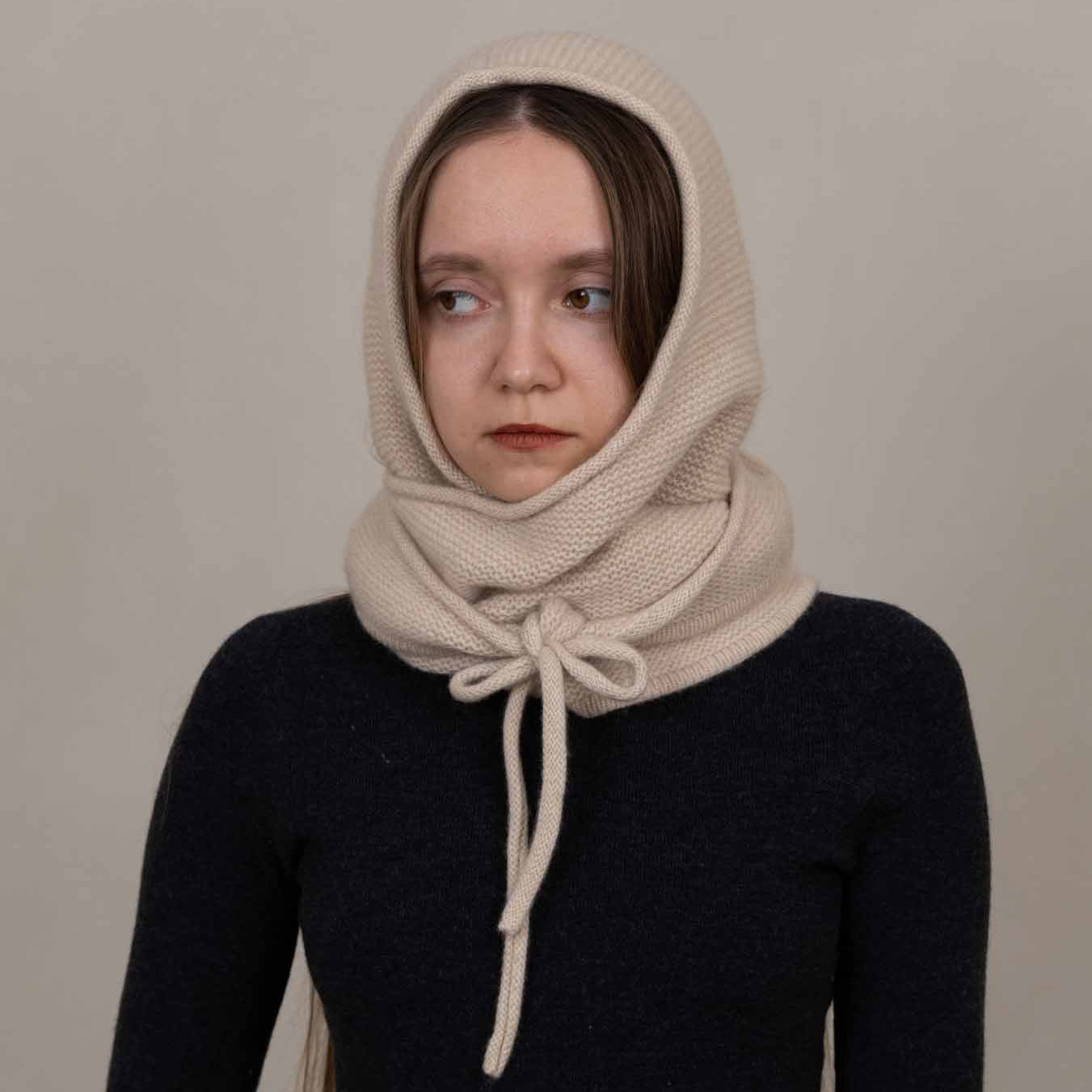 A woman wearing a cream-colored knit head and neck scarf tied at the front, paired with a dark navy long-sleeve top, against a plain light gray background.