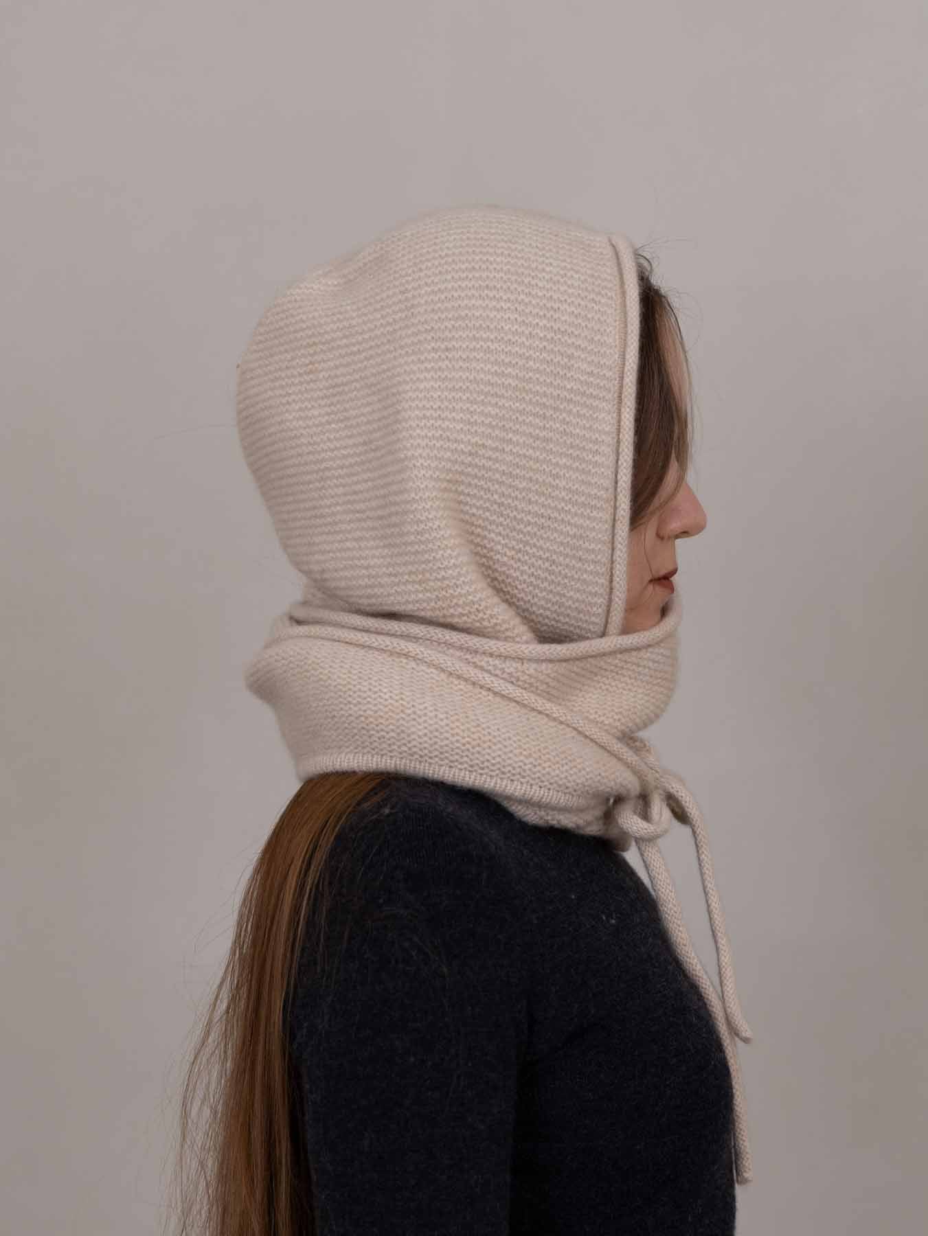 A side-profile view of a person wearing a cream-colored ribbed knit bonnet and matching neck gaiter against a plain light gray background.