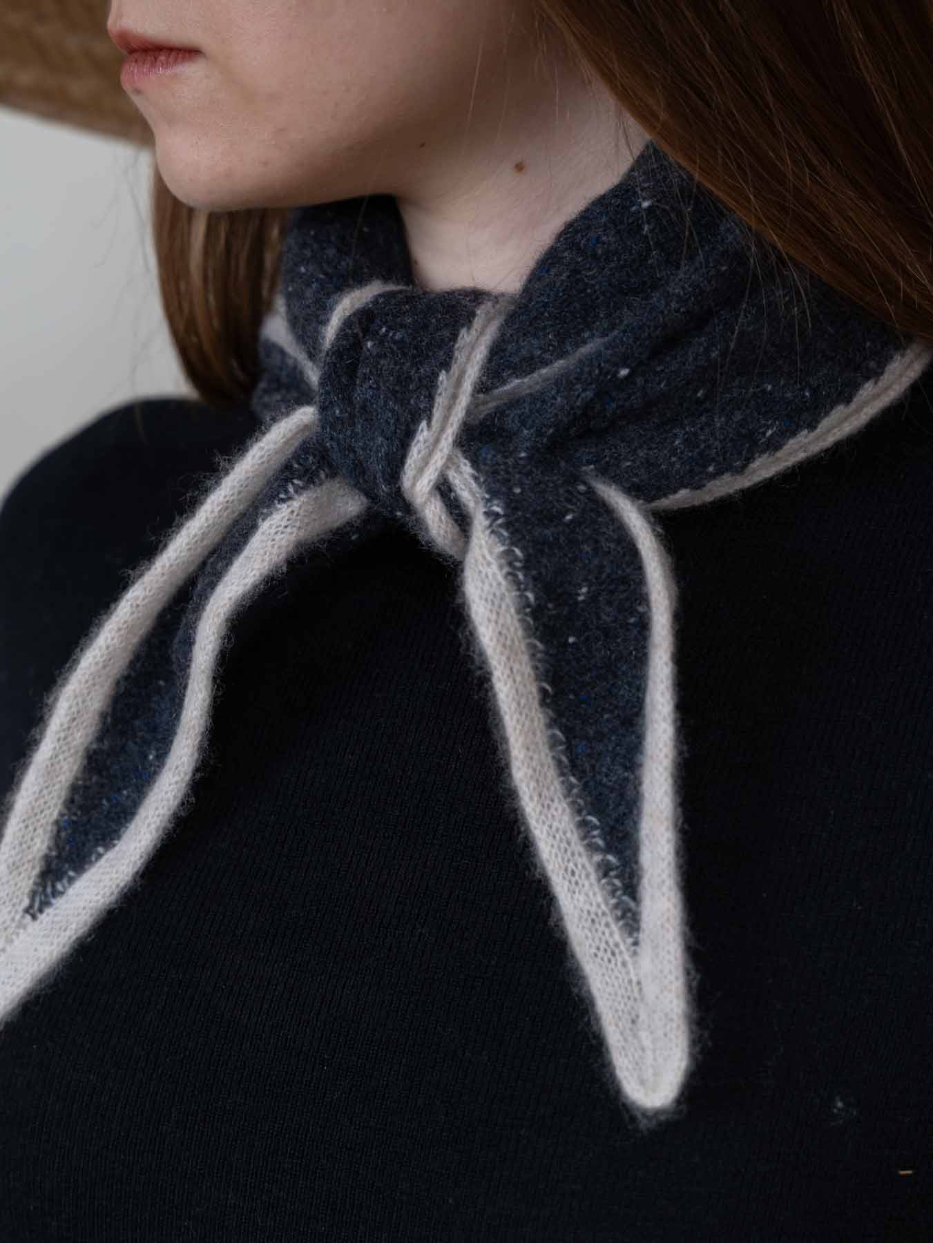 Close-up of a person wearing a dark navy knit scarf tied around the neck over a black top, with light cream trim and a soft textured finish.