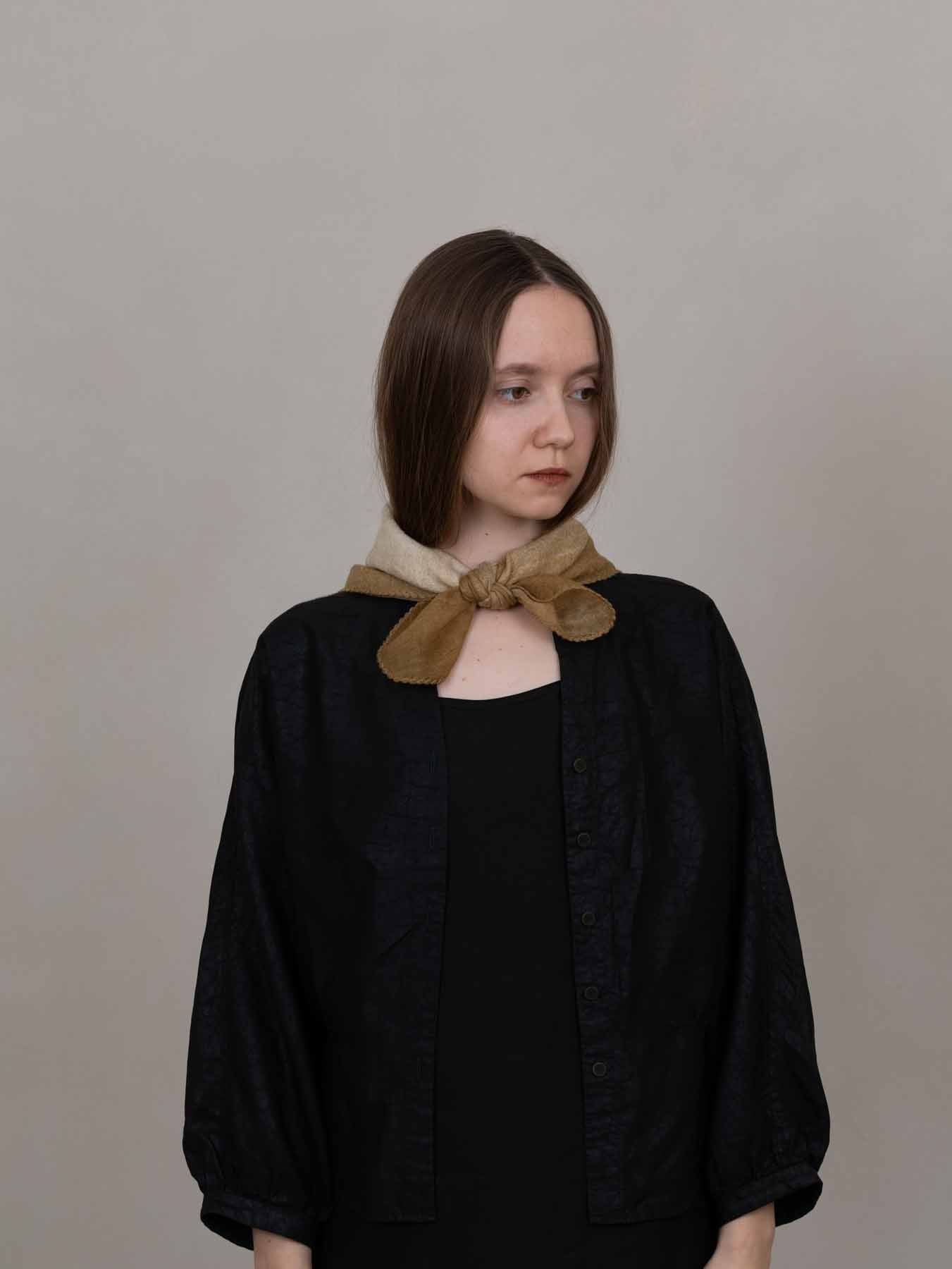 A model with shoulder-length brown hair wearing a black textured jacket over a black top, styled with a wide tan scarf tied in a bow at the collar, set against a plain light gray background.