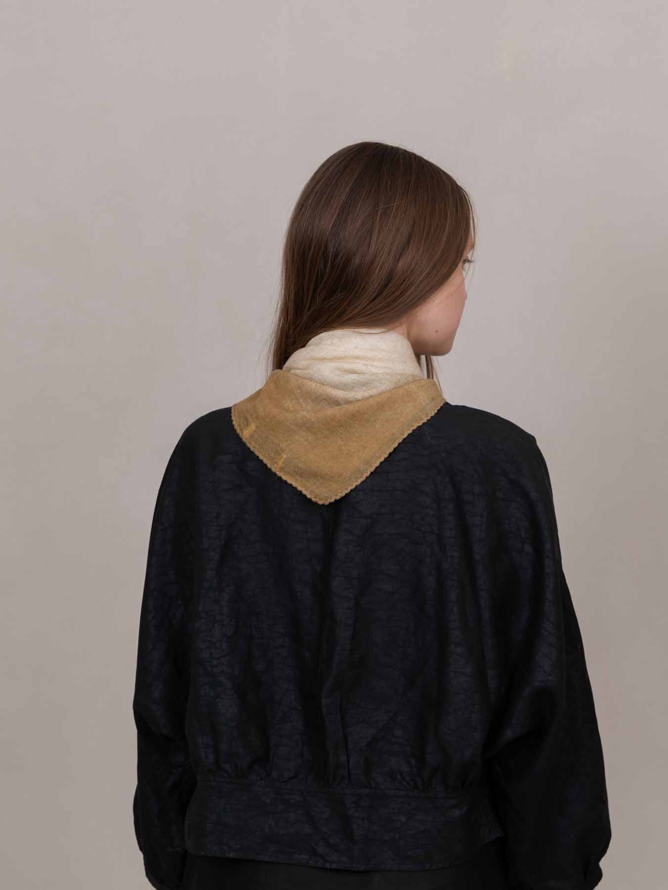 A woman viewed from behind wearing a dark navy textured jacket with a contrasting tan collar and a cream neck warmer, set against a plain light gray background.