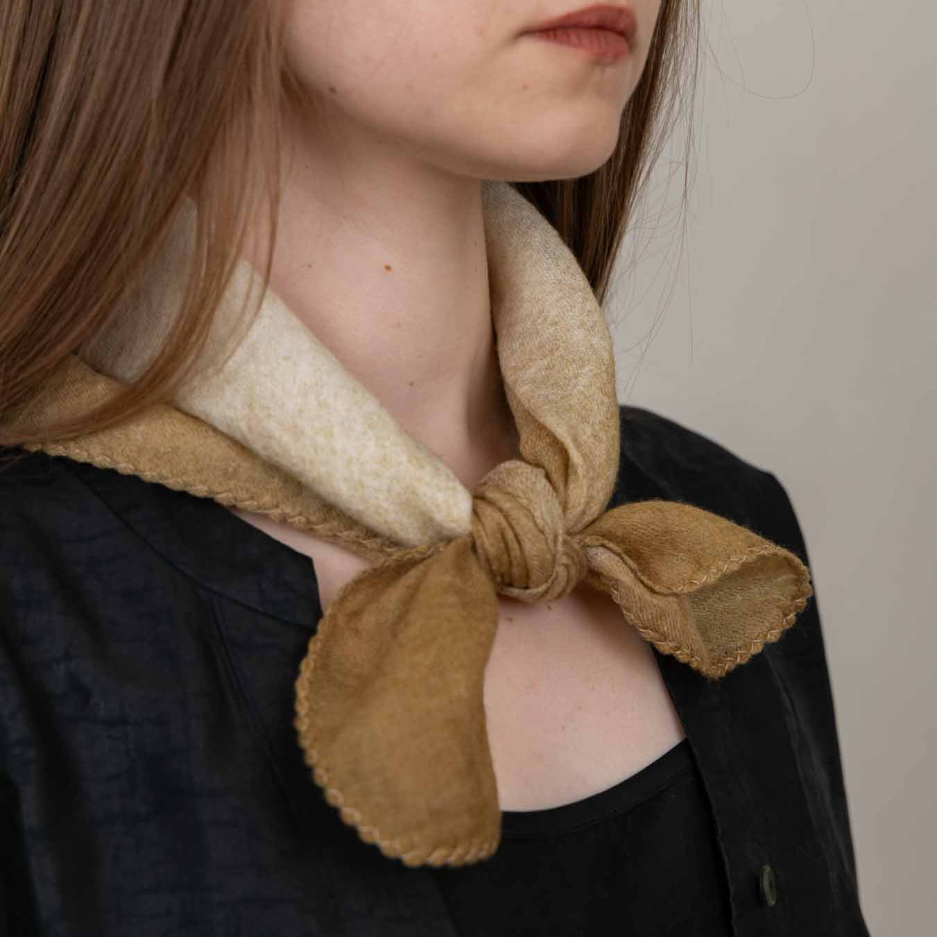 A close-up of a model wearing a two-tone scarf with a cream inner lining and a tan suede-like exterior, tied in a knot at the collar over a black top.