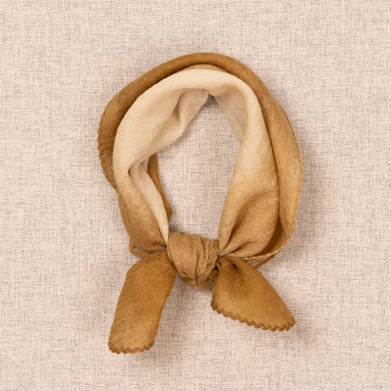 A soft fabric headband or hair scarf tied in a knot, shown flat on a textured beige linen-like background. The outer layer is a warm camel brown, and the inner layer is a lighter cream tone.