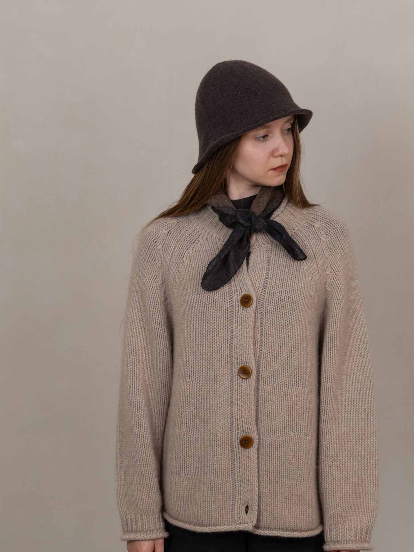 Person wearing a beige knitted cardigan with brown buttons, a dark hat, and a black scarf against a plain background.