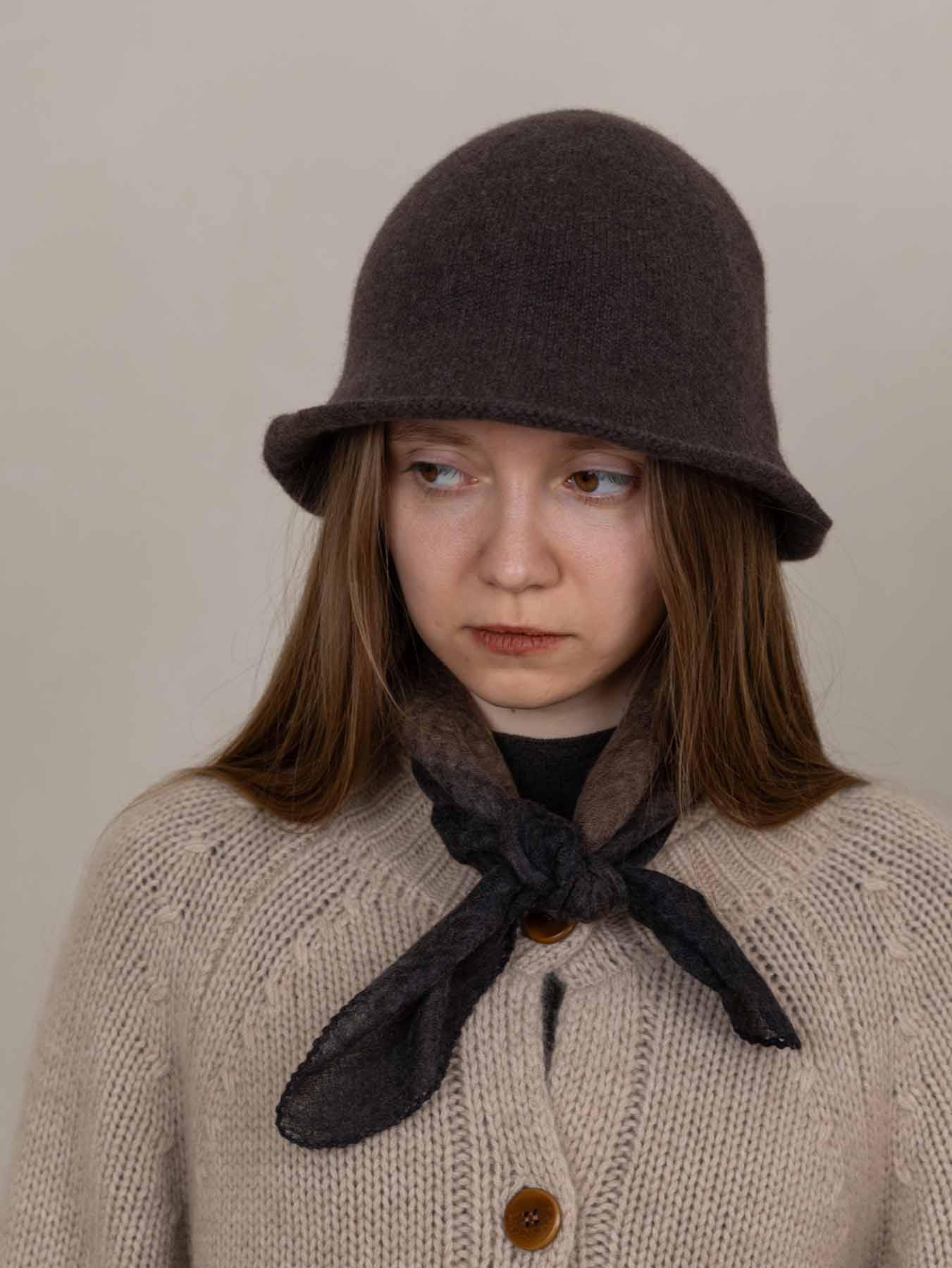 Person wearing a brown knitted hat and beige sweater with a dark scarf against a plain background