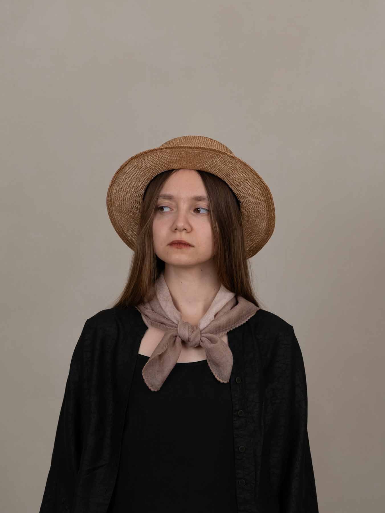 Woman wearing a straw hat and black outfit with a beige scarf against a plain background