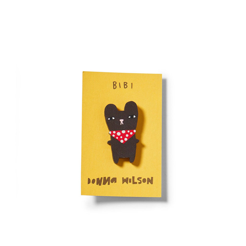 A black brooch shaped like a bear wearing a red polka dot bandana on a yellow card. Brand: Donna Wilson.