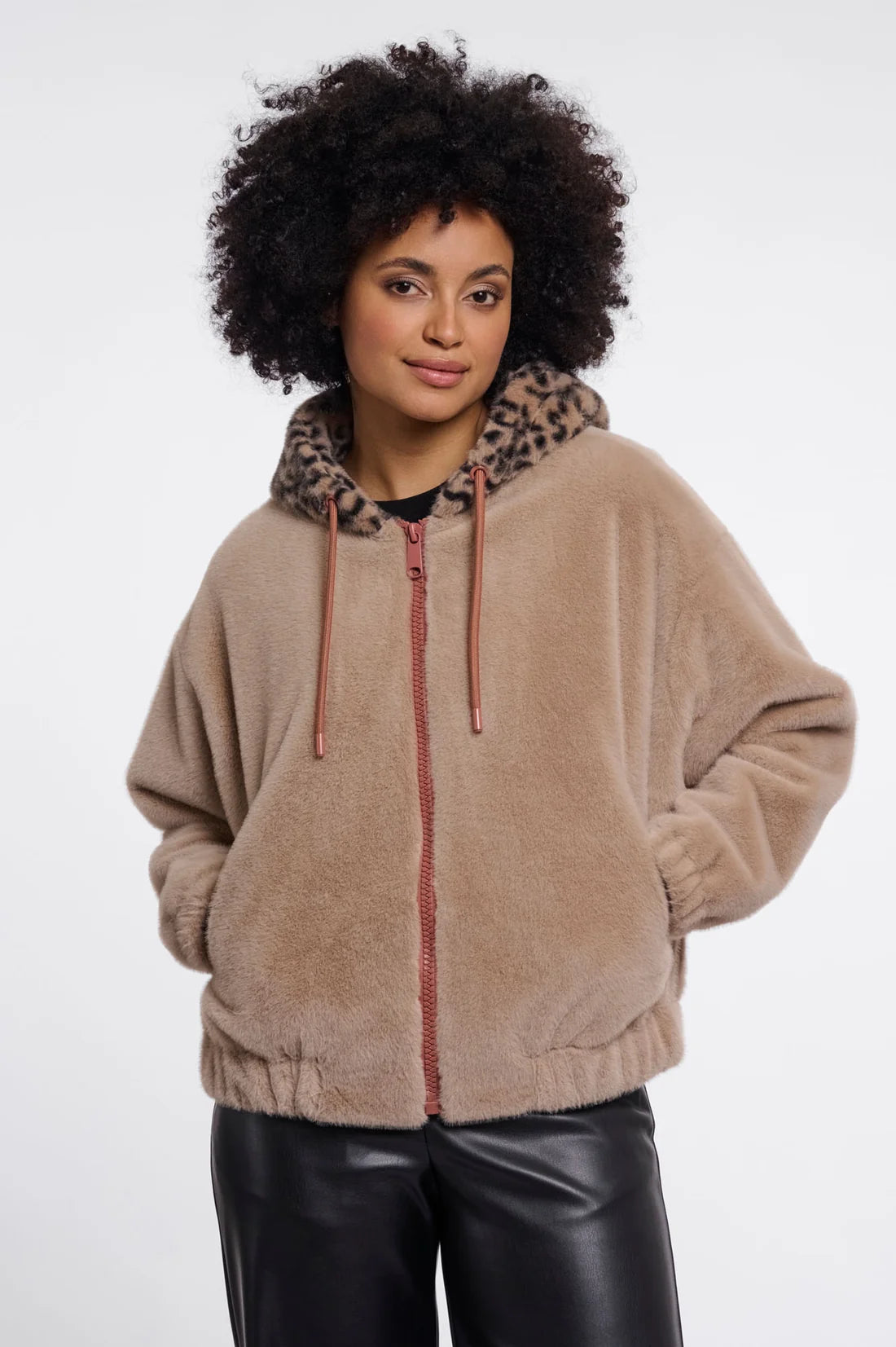 A woman with an afro hairstyle wearing a beige sherpa zip-up hoodie with a leopard-print hood lining and black leather pants, photographed against a plain white studio background.