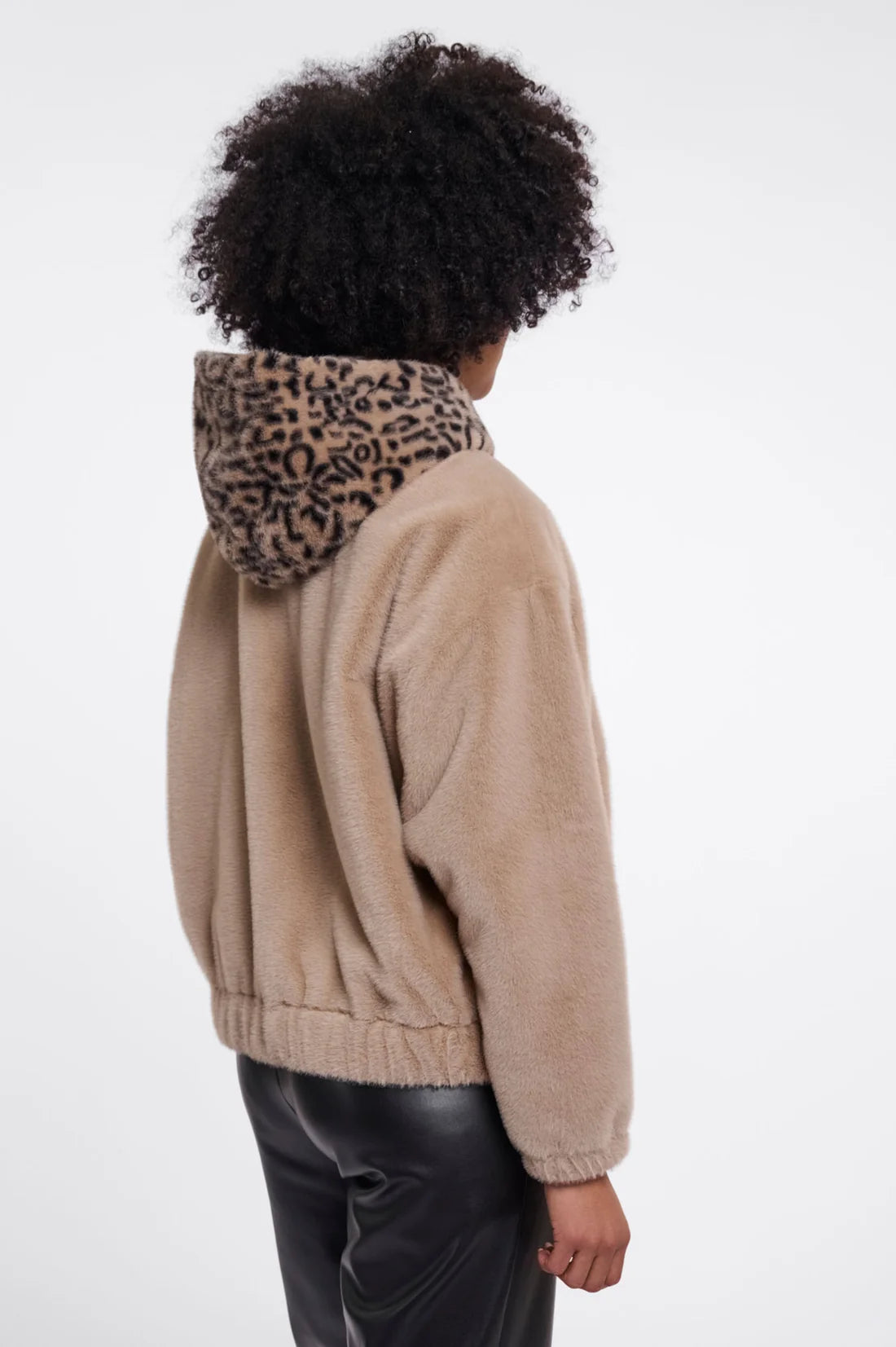 A child shown from the back wearing a beige fuzzy hooded jacket with a leopard-print hood and black pants, photographed against a plain light gray studio background.