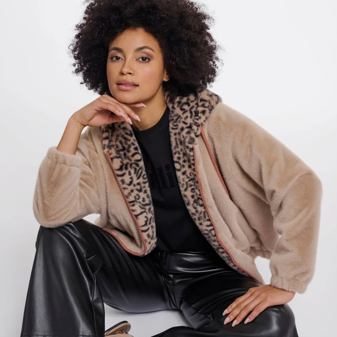 A model with an afro sits on a white studio floor wearing a beige sherpa jacket with a leopard print lining, black leather pants, and leopard print socks.