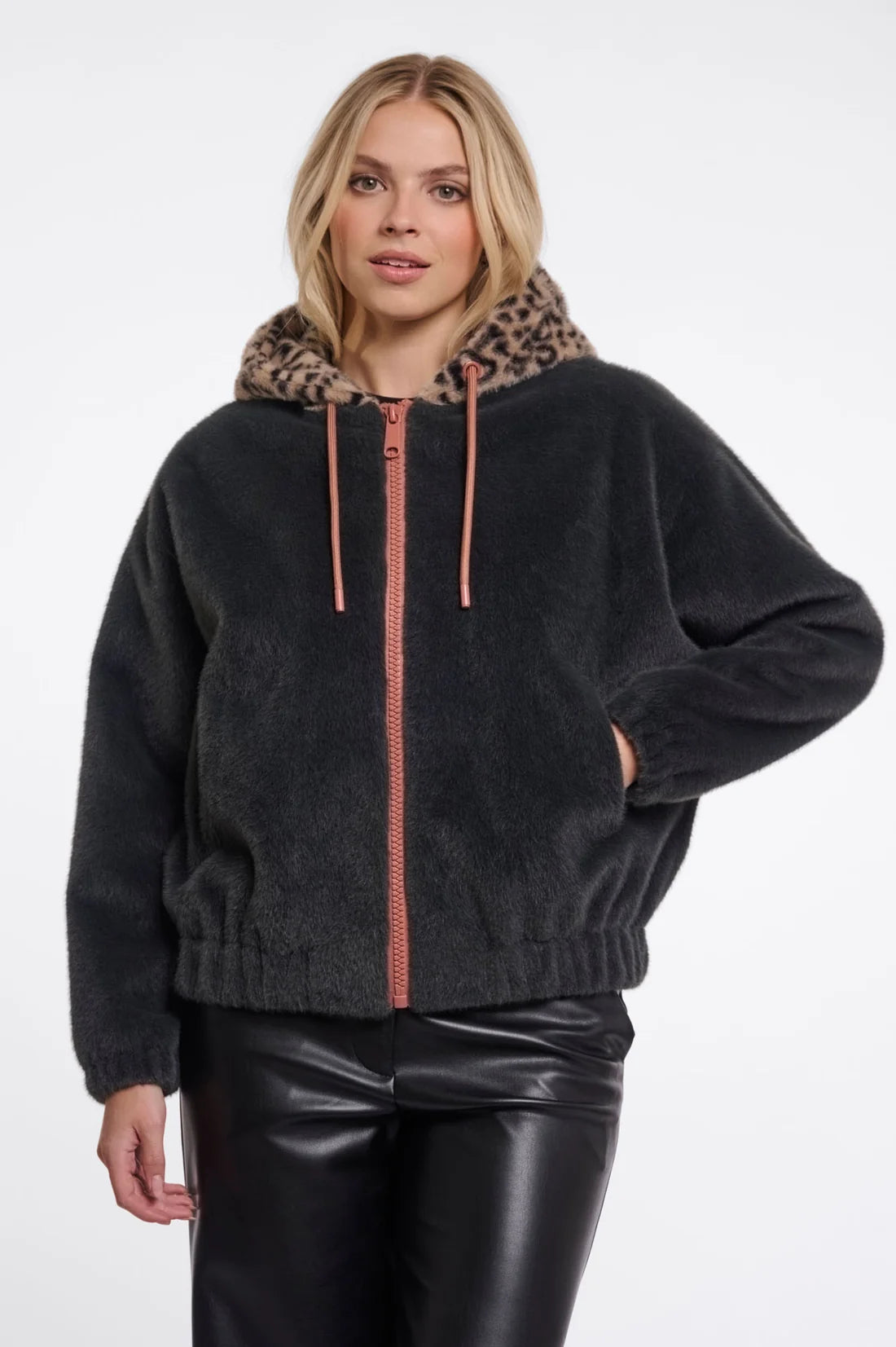 A woman wearing a dark charcoal fuzzy zip-up hoodie with a leopard-print hood lining and pinkish drawstrings, paired with black faux-leather pants, against a plain white studio background.