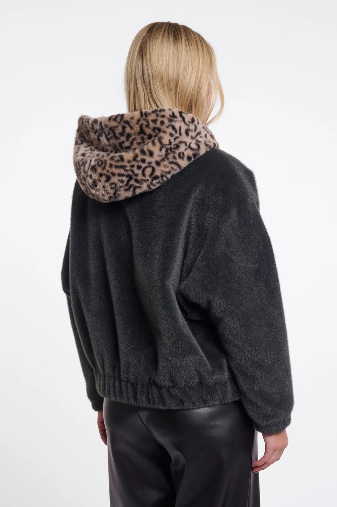 A model shown from the back wearing a dark charcoal fuzzy cropped hoodie with a leopard-print hood, paired with black leather pants, against a plain white studio background.