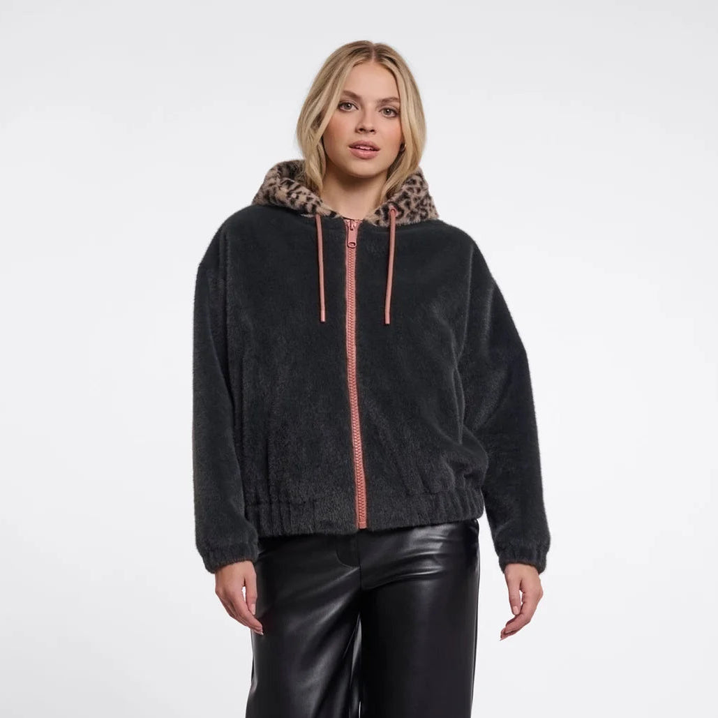 A woman modeling a black fuzzy zip-up hoodie with a leopard-print hood lining and pink drawstrings, paired with black faux-leather pants, against a light gray studio background.