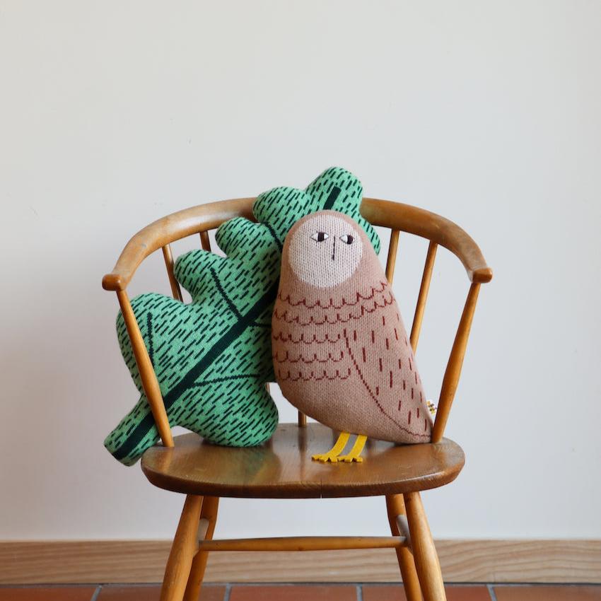 Two colorful cushions by Donna Wilson: a brown owl with yellow feet and a green leaf with black details on a wooden chair.