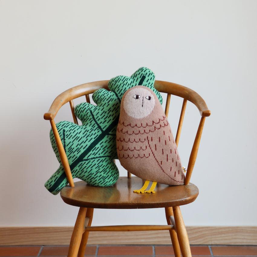 A wooden chair holds a brown owl pillow with yellow feet and an oak leaf pillow in green; both by Donna Wilson.