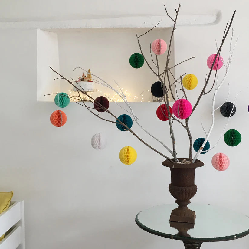 Colorful honeycomb paper ball ornaments hanging from a bare branch in a dark urn on a glass-topped table.