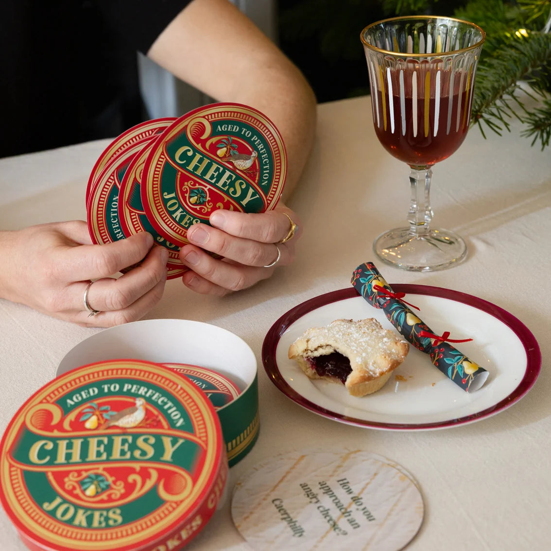 Person holding 'Cheesy Jokes' coasters with a festive setting including a glass of red drink and a cracker.