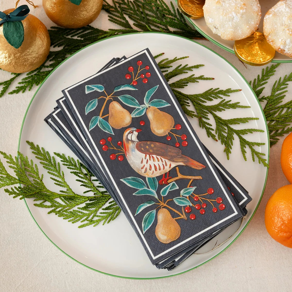 Stack of decorative napkins with bird and pear design on a plate with greenery and oranges.