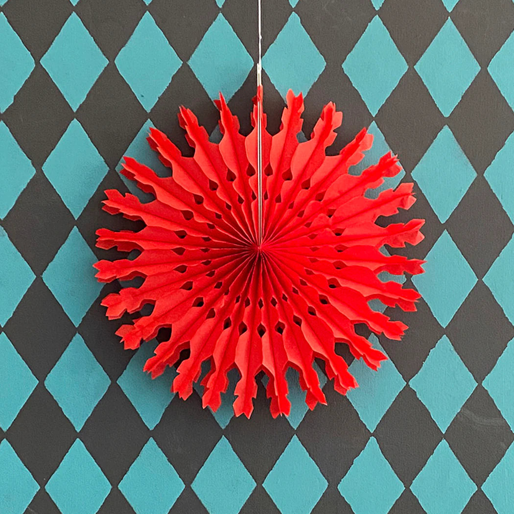 Red paper snowflake decoration with a lacy design, hanging against a teal and black diamond pattern background.