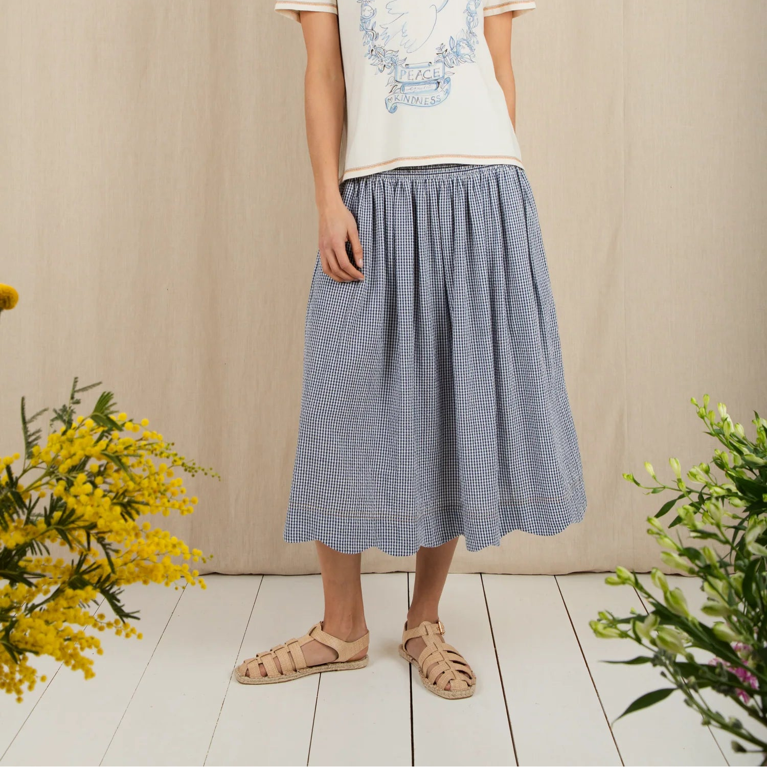 Woman wearing a white t-shirt with a graphic design and a blue skirt, standing against a beige wall with plants.
