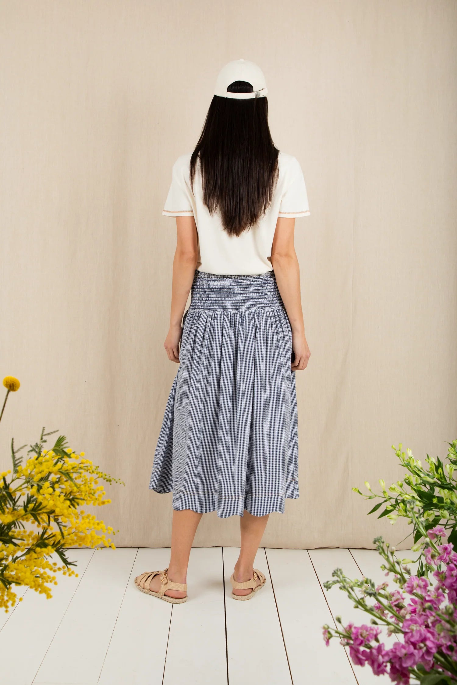Person wearing a white t-shirt and blue skirt standing in front of a beige wall with flowers on either side.