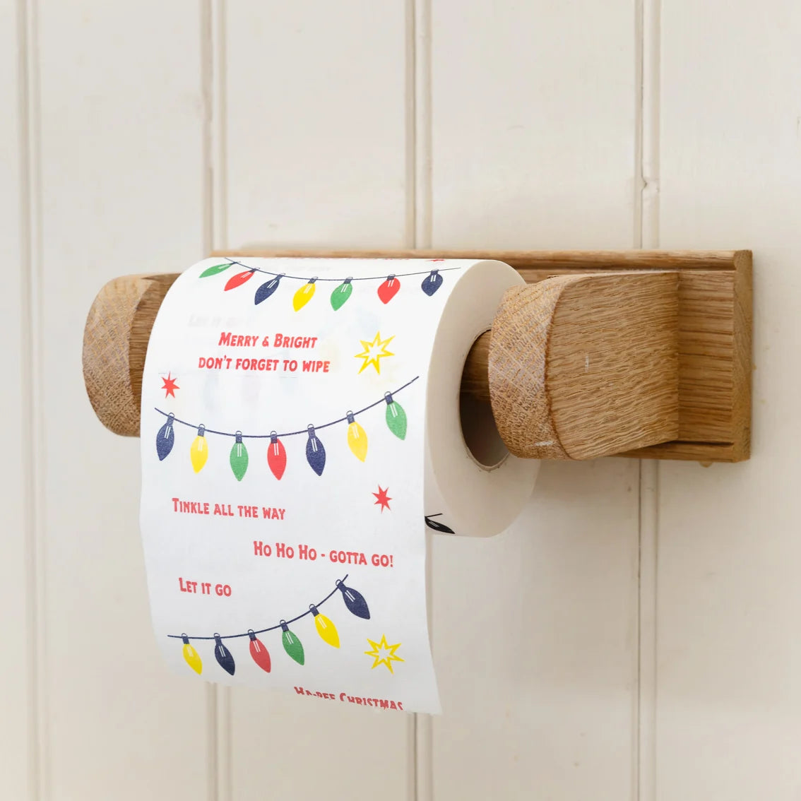 A roll of toilet paper with a Christmas-themed print featuring colorful string lights and festive quotes, mounted on a wooden holder against a white paneled wall.