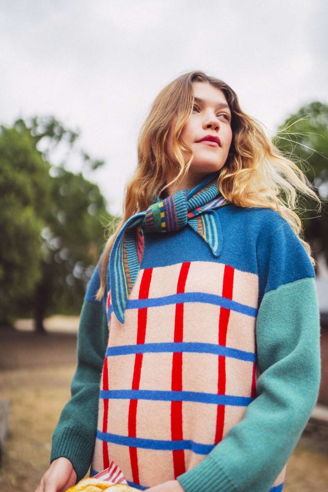 A woman wearing a colorful knit sweater with a blue and green color-blocked design and a red-and-beige grid pattern on the front, photographed outdoors in a park.