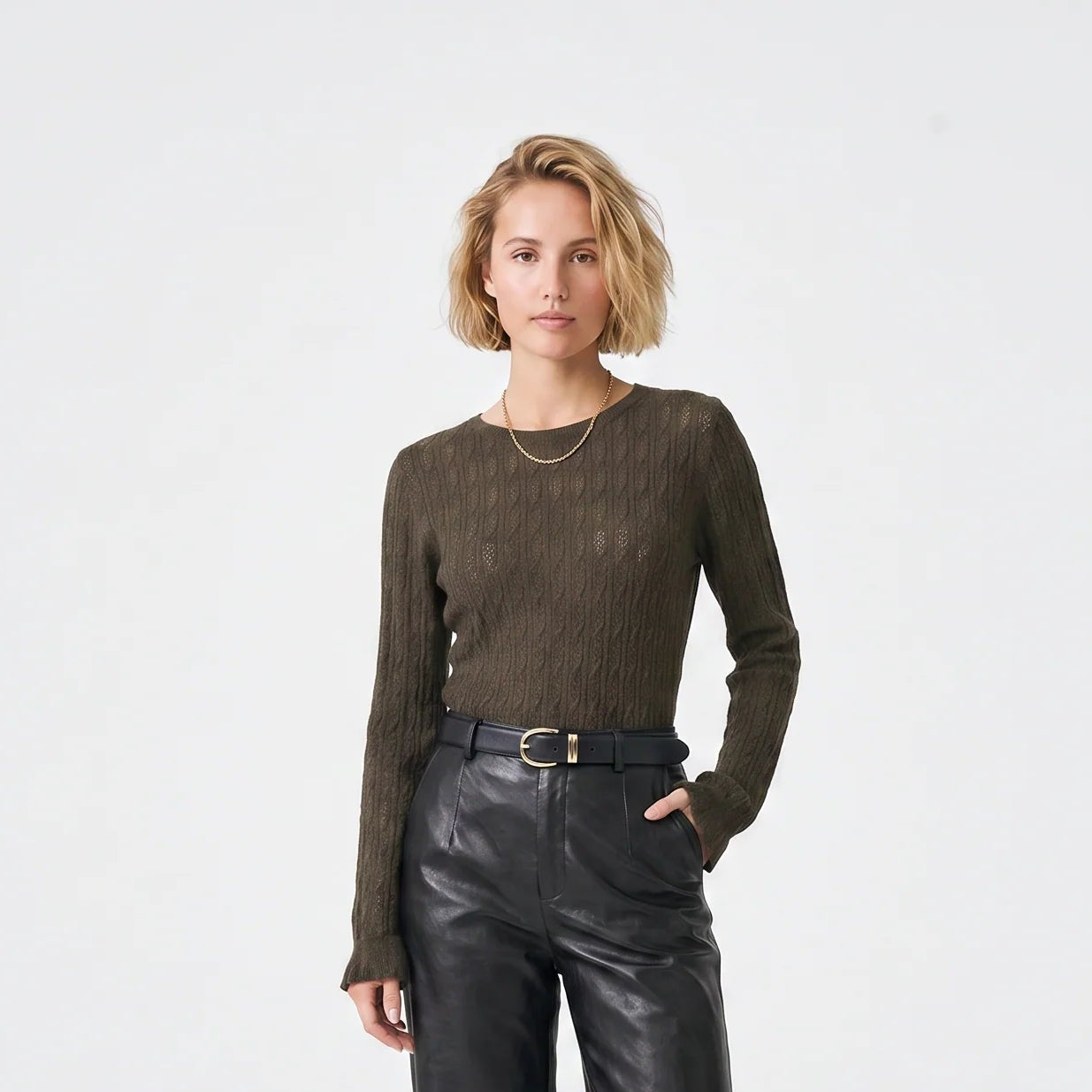 A model stands against a light gray studio background wearing a dark olive-brown ribbed long-sleeve top, black wide-leg leather-look pants, a black belt, and black flats.