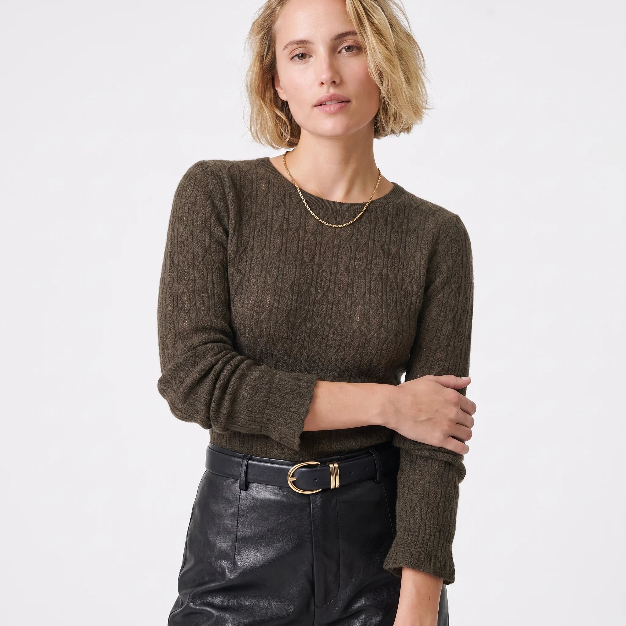 A woman wearing a dark brown cable-knit crewneck sweater and black leather pants with a gold buckle belt, photographed against a clean white studio background.