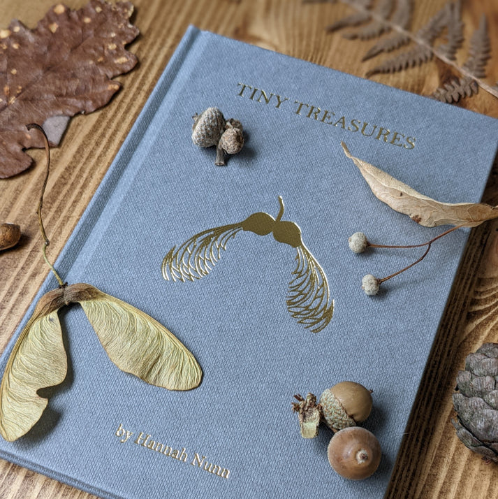Gray linen-textured book titled "Tiny Treasures" by Hannah Nunn; gold-foil seed illustration on cover.