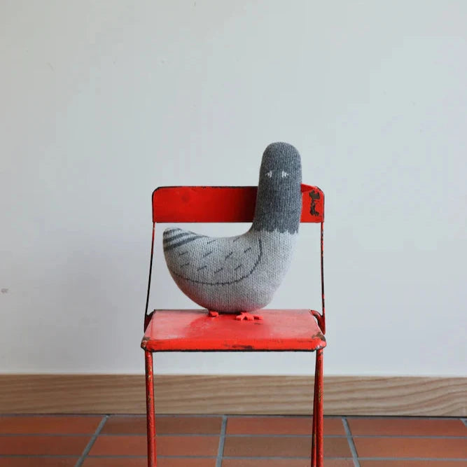 A grey knitted pigeon by Donna Wilson sits atop a red metal chair against a neutral background.