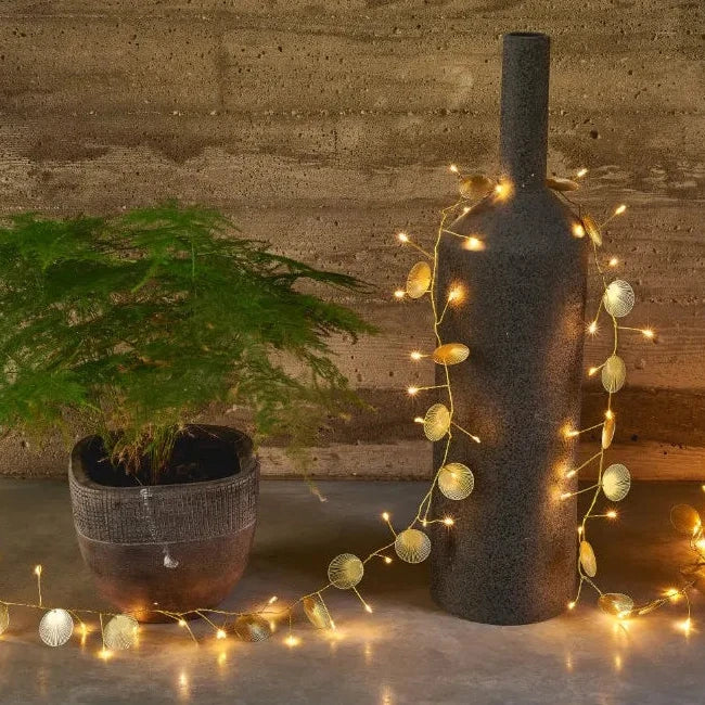 Gold leaf-shaped LED string lights draped around a dark vase.  Warm-toned lights illuminate the metallic accents.