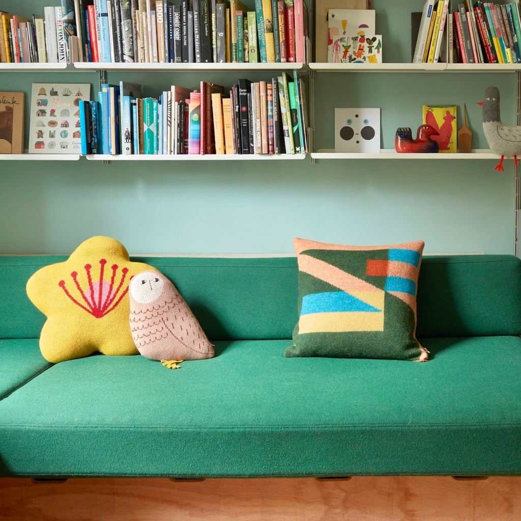 A green sofa with a yellow flower cushion, a brown owl cushion, and a patterned Donna Wilson pillow on it.